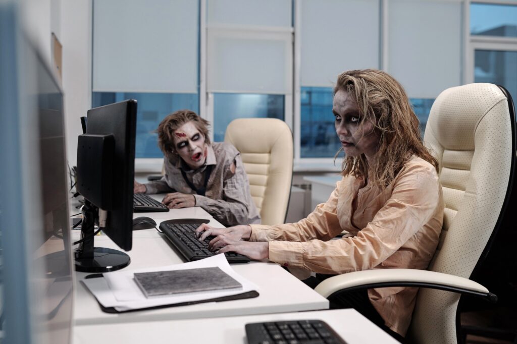 Young spooky zombie businesswoman sitting by desk in front of computer monitor and networking